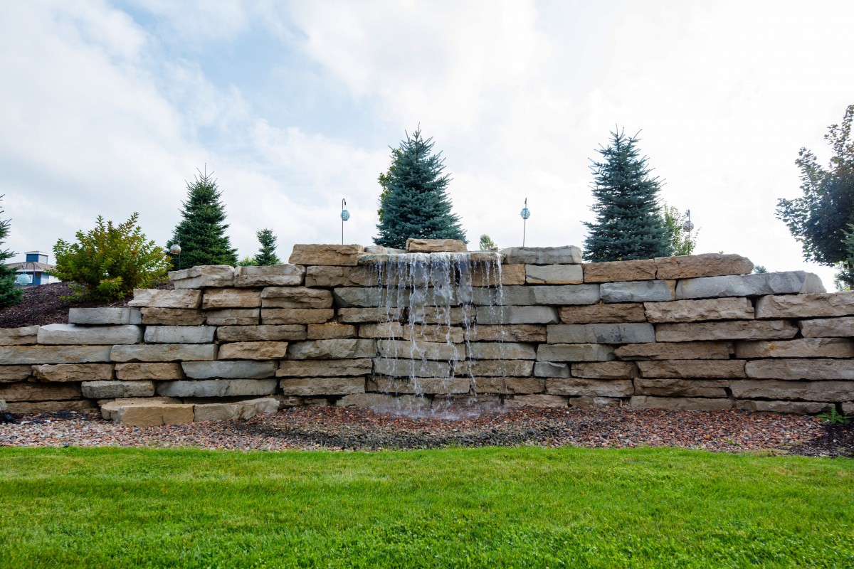 A small waterfall cascading over a stone wall in a landscaped garden setting.
