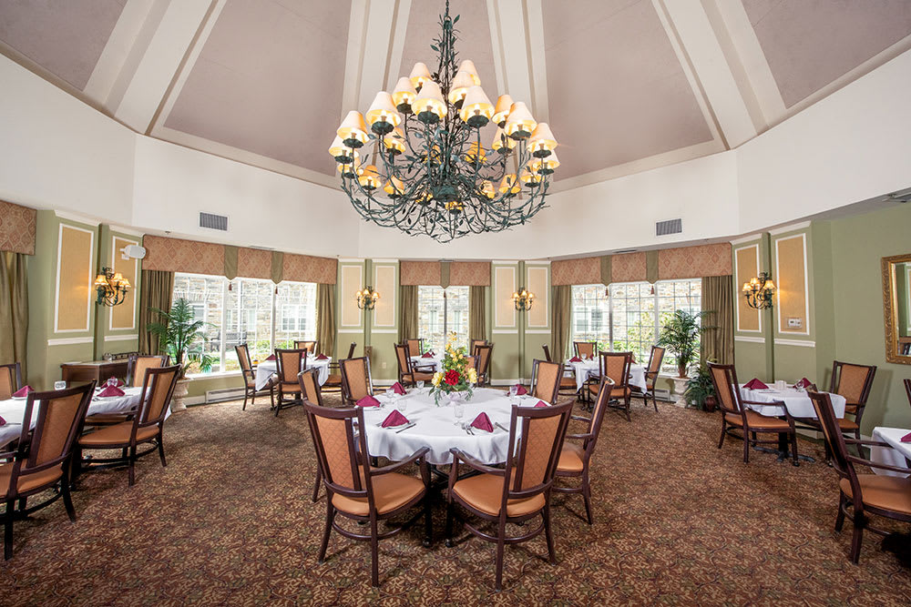An elegant dining room with a large chandelier, neatly arranged tables and chairs, and large windows offering natural light.