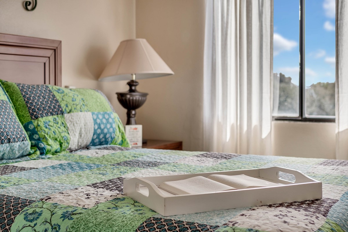 A cozy bedroom with a quilted bedspread and a decorative lamp, illuminated by natural light from the window.