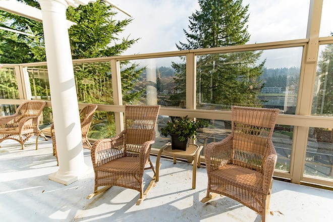 A peaceful patio setup with wicker chairs, surrounded by glass railings and greenery, on a sunny day.