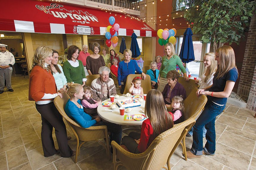 A family celebrates a birthday with children gathered around a cake at an indoor party, surrounded by balloons and decorations.