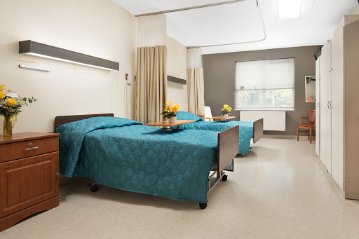 A well-lit hospital patient room featuring two beds, each adorned with teal bedspreads and a small vase of flowers.
