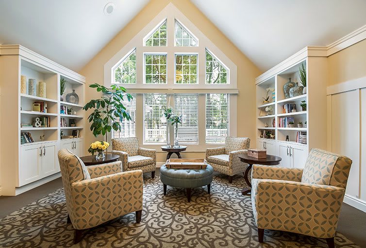 A cozy living room with armchairs arranged around a coffee table, natural light coming through large windows and bookshelves lining the walls.