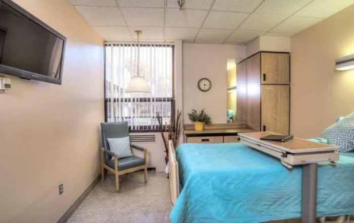 A tidy hospital room with a bed, chair, and window featuring vertical blinds.