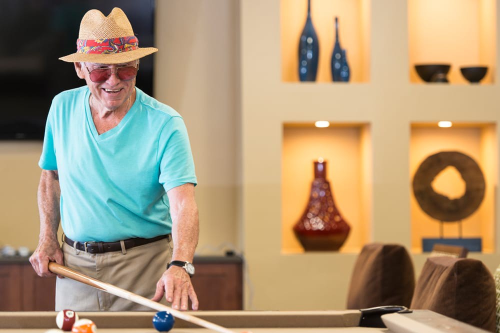 An elderly man wearing a hat enjoys playing billiards indoors.