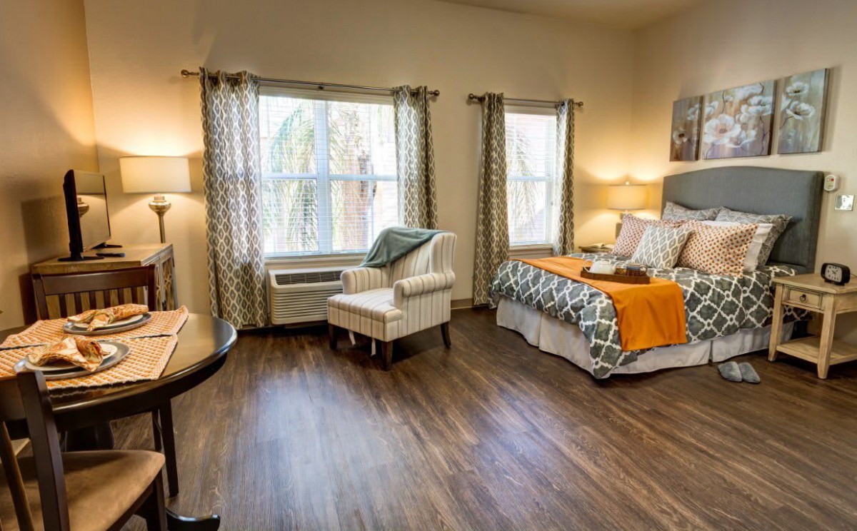 A cozy bedroom with wood flooring, featuring a bed adorned with decorative pillows and a small armchair by the window that allows plenty of natural light.