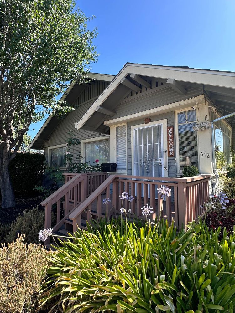 A charming house with a porch surrounded by a garden and trees on a sunny day.