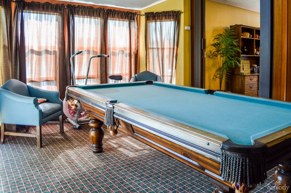 A cozy game room featuring a classic pool table, surrounded by large windows with drapes, and comfortable seating on a patterned carpet.
