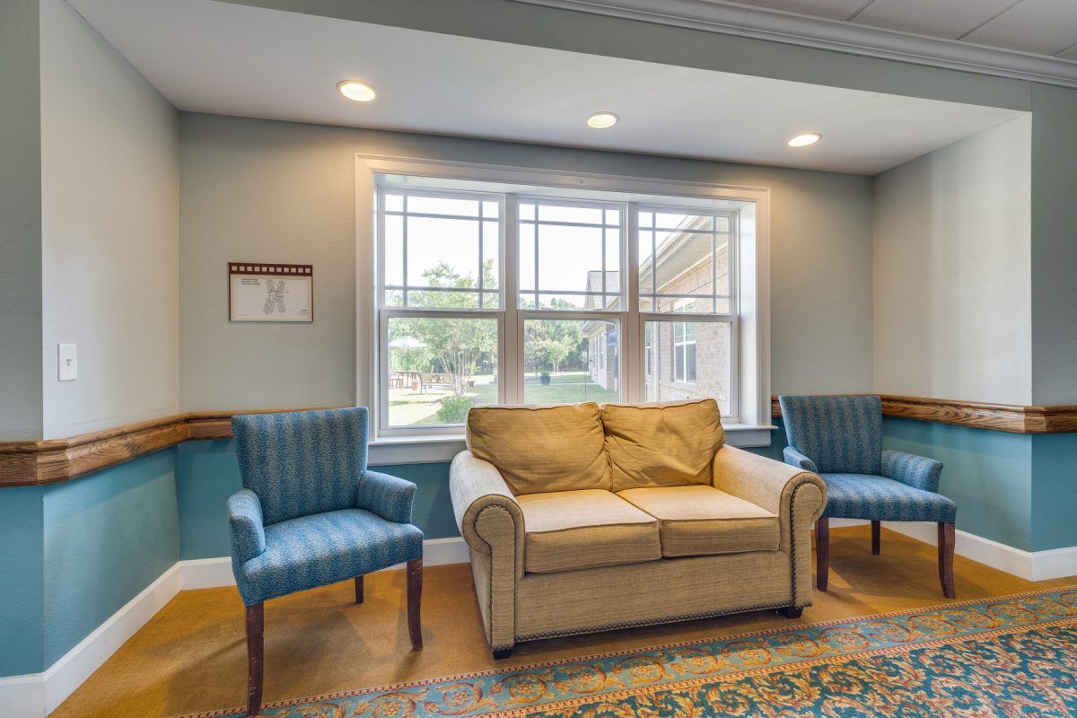 A cozy living room with a beige couch, two blue chairs, and a large window letting in natural light.
