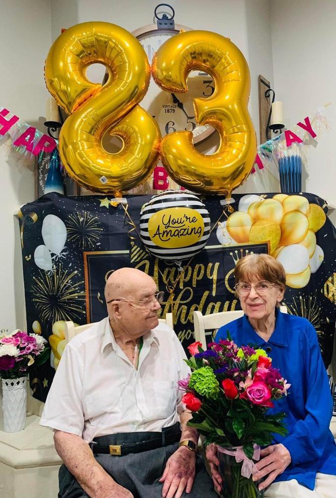 An elderly couple celebrating a birthday with balloons and flowers.