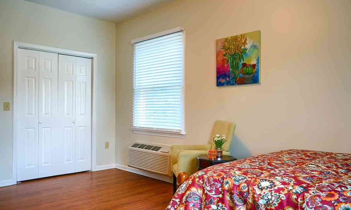 A cozy bedroom with a colorful bedspread, an armchair by the window, and artwork on the wall.