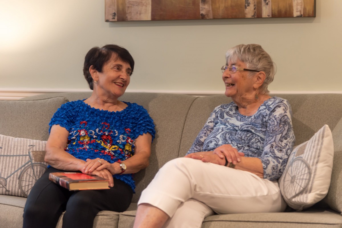 Two elderly women sitting on a couch, smiling and talking to each other.