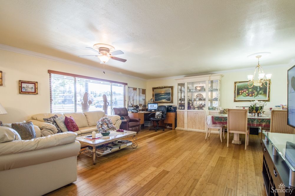 A spacious living room with wooden flooring, featuring a comfortable sofa set, coffee table, and large windows allowing natural light.