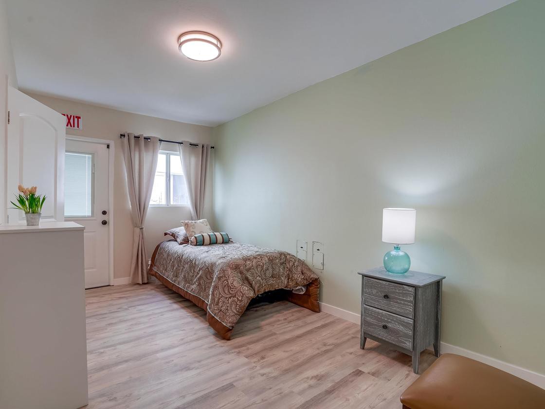 A cozy bedroom with a single bed, nightstand, and lamp, featuring light green walls and hardwood flooring.
