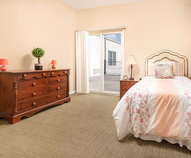 A cozy bedroom featuring a bed with floral bedding, a wooden dresser, and a lamp by the window.