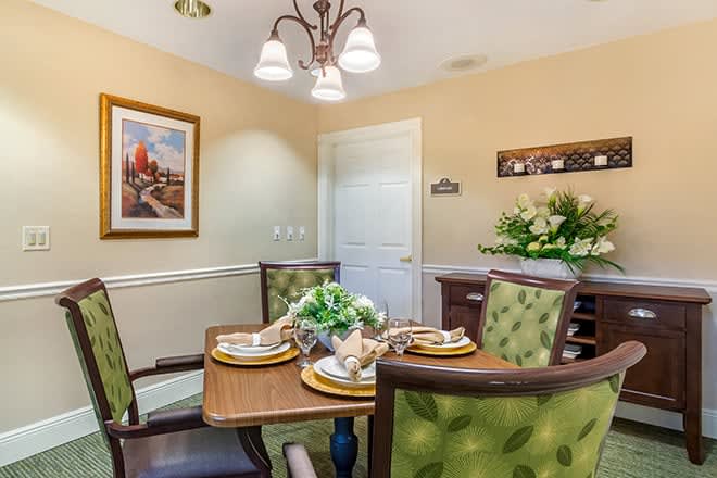 A cozy dining room with a wooden table, green cushioned chairs, and elegant table settings. The decor includes a vibrant plant arrangement and framed artwork on the wall.