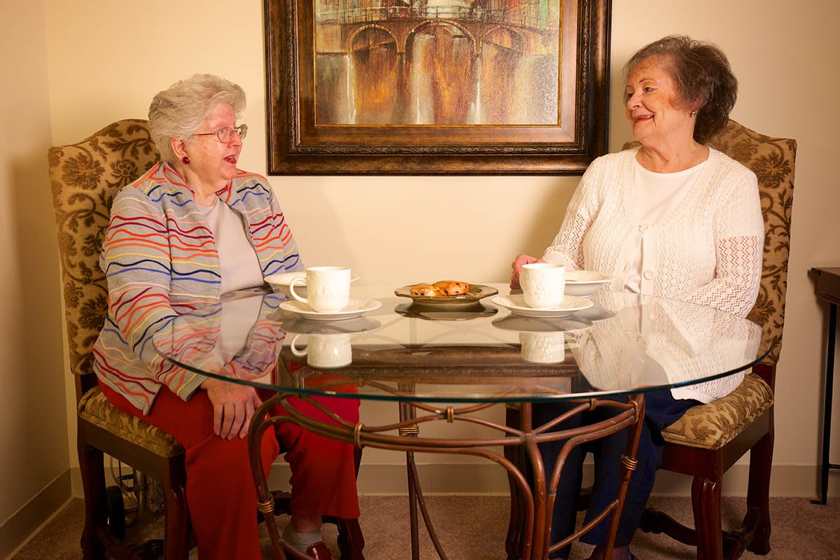 Two elderly women enjoying coffee and conversation at a glass table indoors.