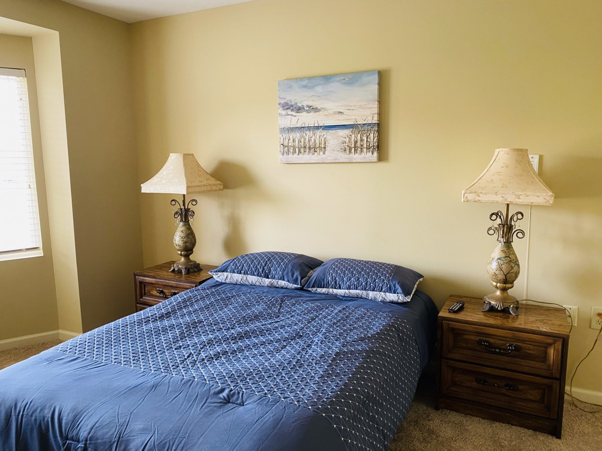 A bedroom with a neatly made bed featuring blue bedding, flanked by two lamps on nightstands, and a piece of artwork above the headboard.