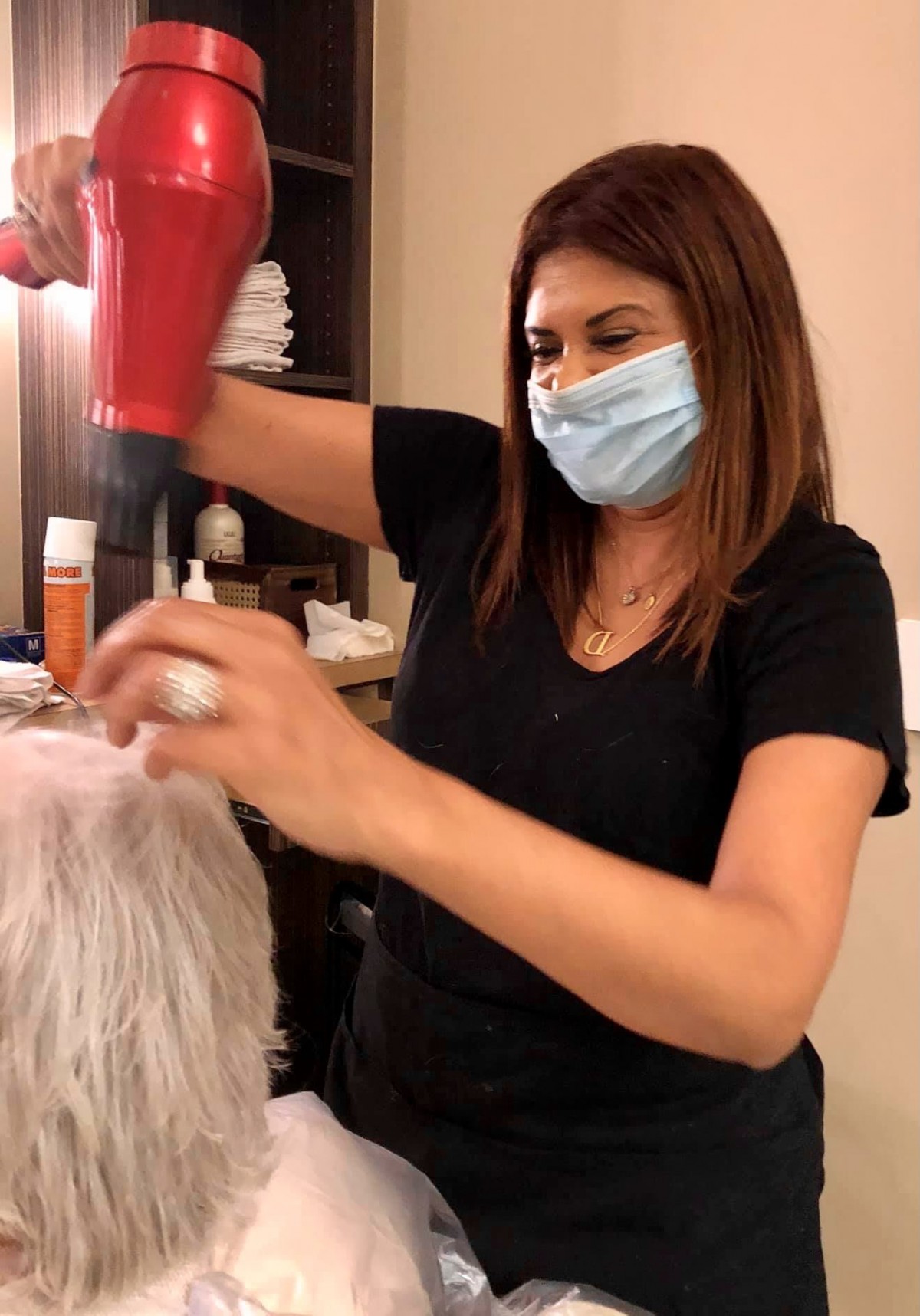 A hairdresser blow-dries a client's hair in a salon while wearing a mask.