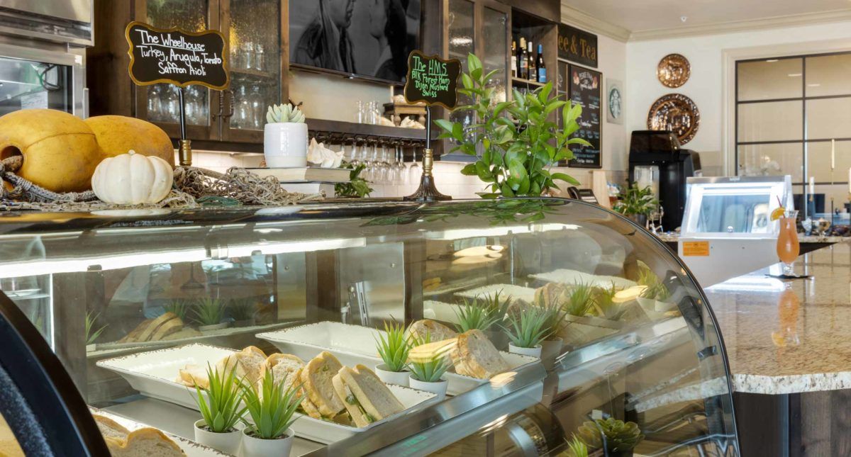 A cozy cafe with a variety of sandwiches displayed in a deli counter, adorned with small potted plants and a menu board in the background.