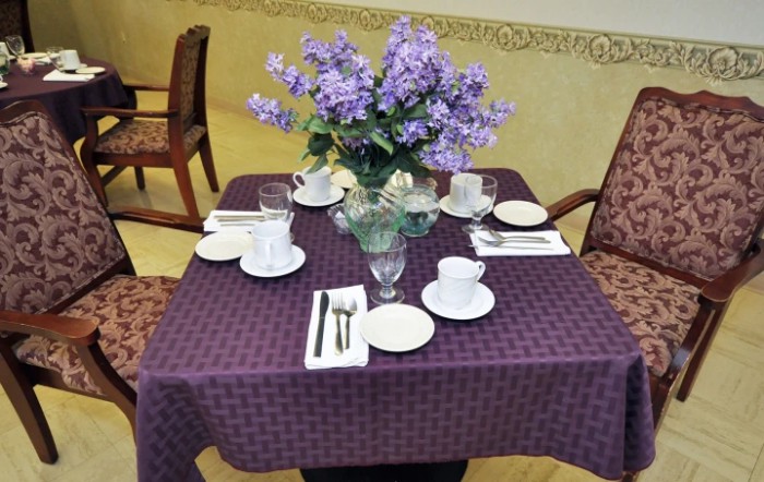 A neatly set dining table with a purple tablecloth and a floral centerpiece, prepared for four people with teacups and cutlery.