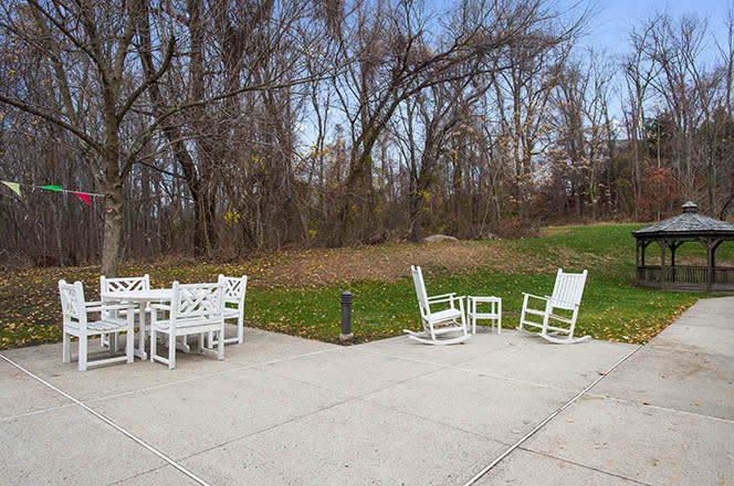A serene outdoor patio area with white furniture, including a table and rocking chairs, bordered by trees and grass.