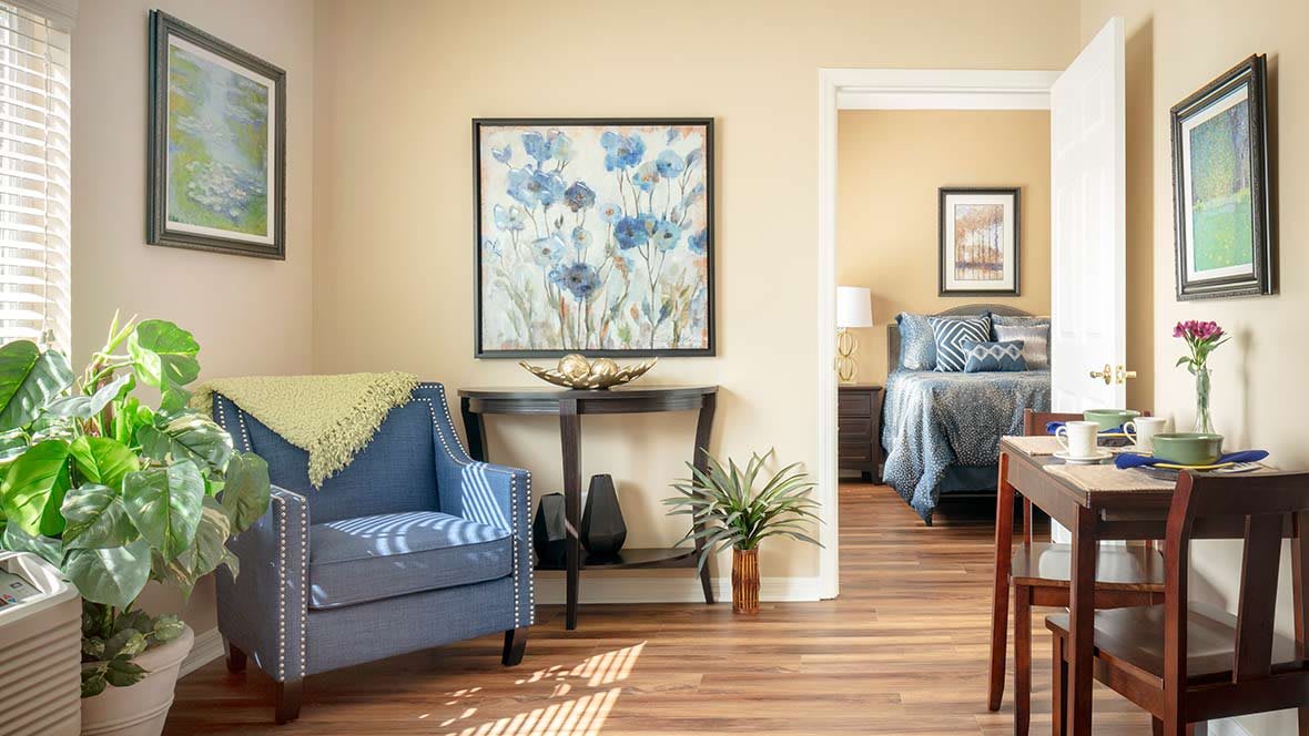A cozy living room with a blue chair and artwork, leading into a bedroom with soft lighting.