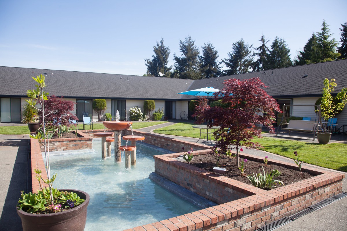 A serene courtyard featuring a central fountain surrounded by lush greenery and brick borders.