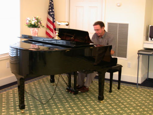 A musician performing on a black grand piano in an indoor setting, accompanied by an American flag.