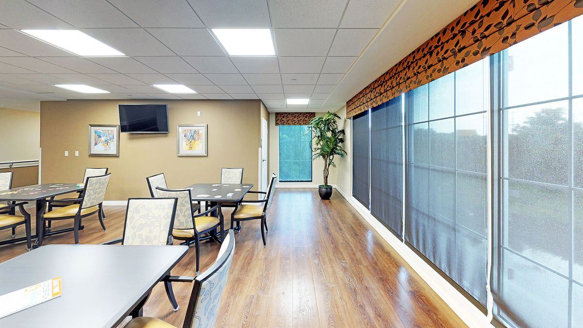 A bright dining area with tables and chairs, featuring wooden flooring, large windows, and decorative plants.