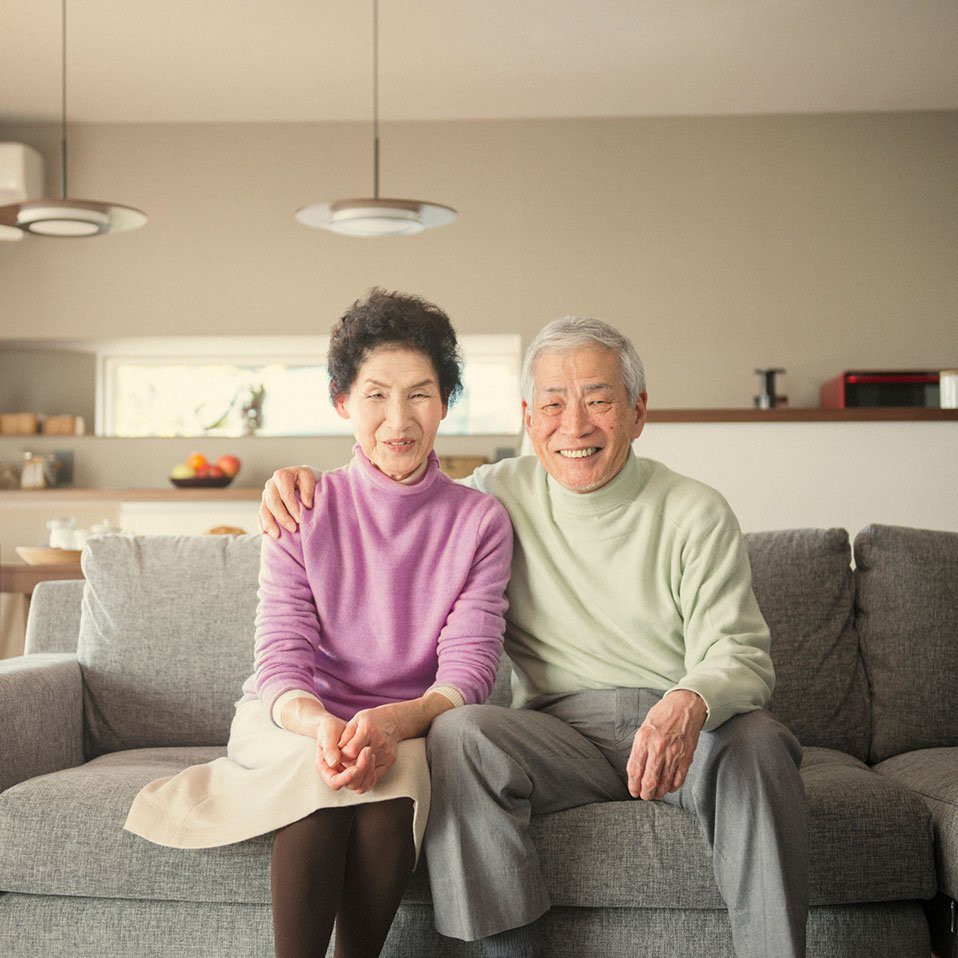 A smiling elderly couple sits together on a sofa in their living room.