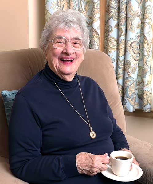 An elderly woman smiles warmly while enjoying a cup of coffee indoors.