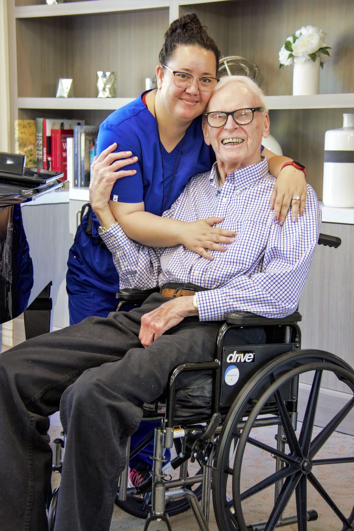 A caregiver affectionately embraces an elderly man in a wheelchair, both smiling happily indoors.