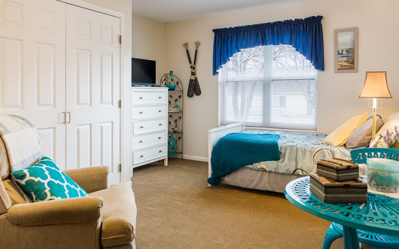 A cozy bedroom featuring a bed with turquoise accents, a white dresser, and a comfortable chair.