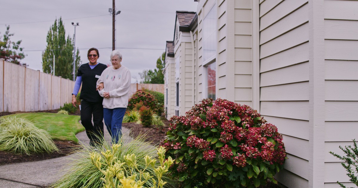A caregiver assists an elderly person on a walk through a garden.