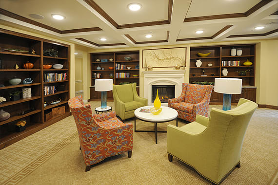 A cozy library reading room featuring armchairs, a coffee table, and bookshelves surrounding a fireplace.