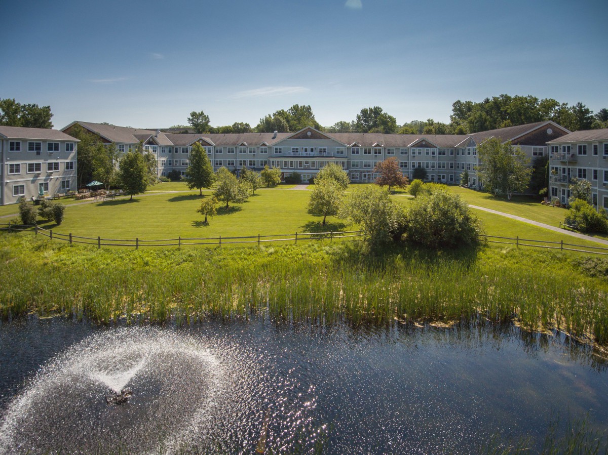 A scenic view of a large building surrounded by greenery with a pond and fountain in the foreground.