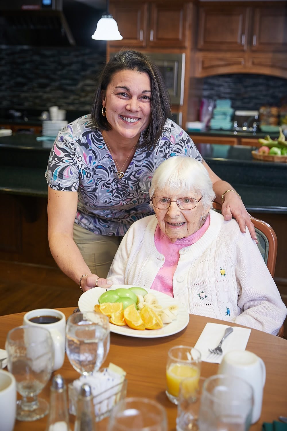 A caregiver smiles as she presents a meal to an elderly woman at the dining table.