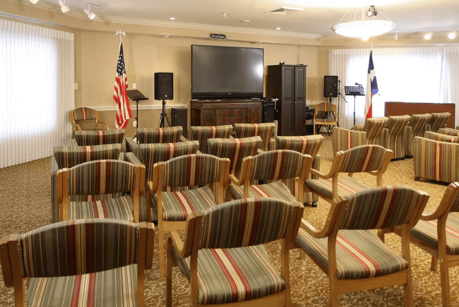 An empty conference room set up with rows of chairs facing a large television screen, flanked by flags.