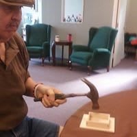 A person is using a hammer on a small wooden object inside a room.