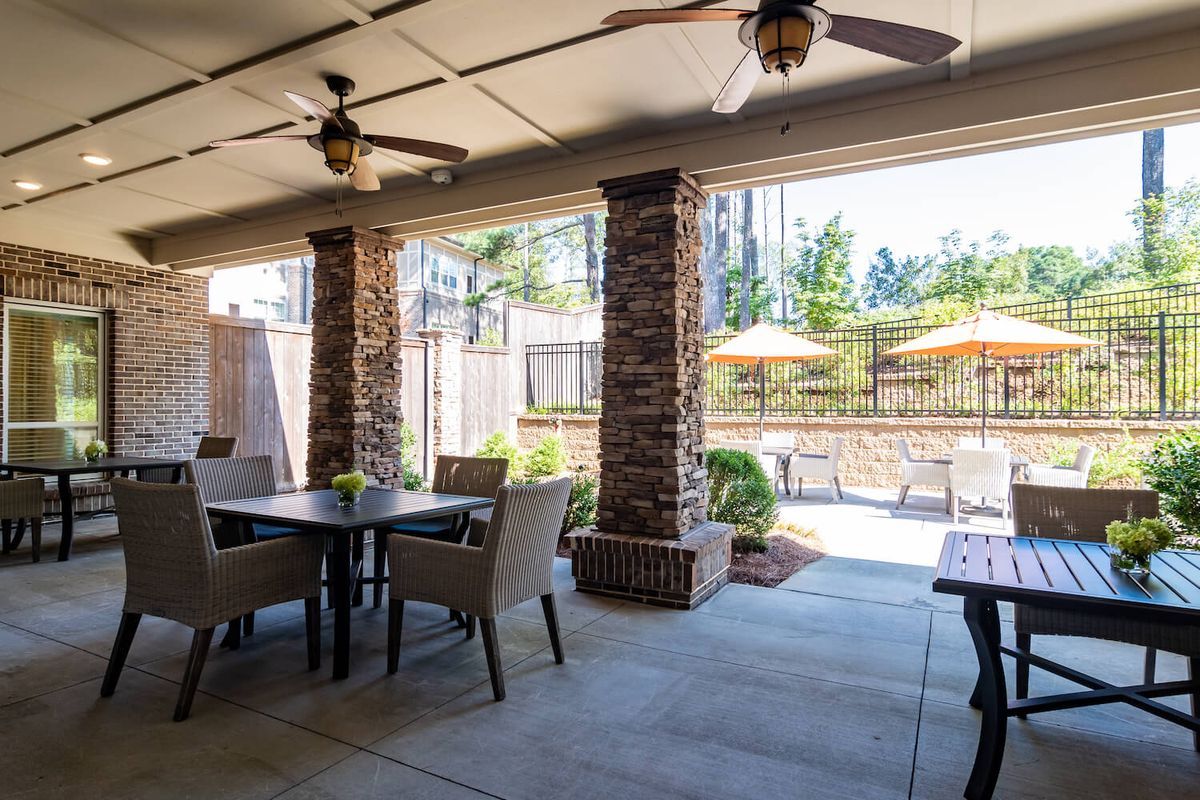 A cozy outdoor patio area with dining furniture and ceiling fans, nestled under a covered structure with brick columns.