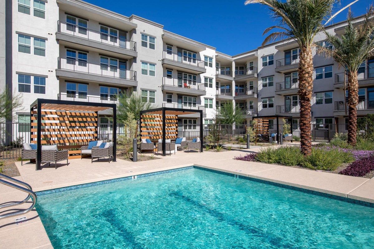 A modern apartment complex with a swimming pool surrounded by outdoor seating and palm trees.