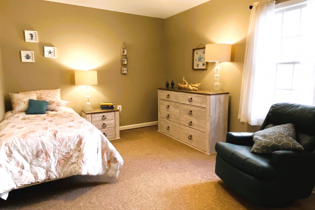 A cozy bedroom with warm lighting, featuring a bed, dresser, and armchair.
