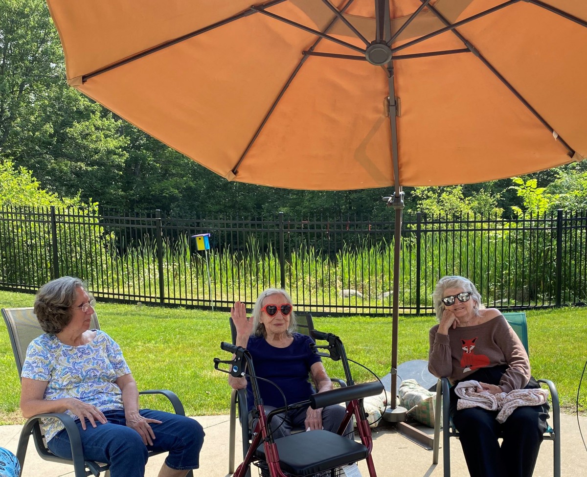 A group of seniors sitting under a large umbrella in a garden on a sunny day.