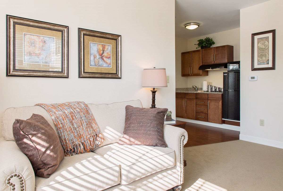 A cozy living room with a sofa and decorative pillows adjacent to a kitchen area, featuring warm lighting and artwork on the walls.
