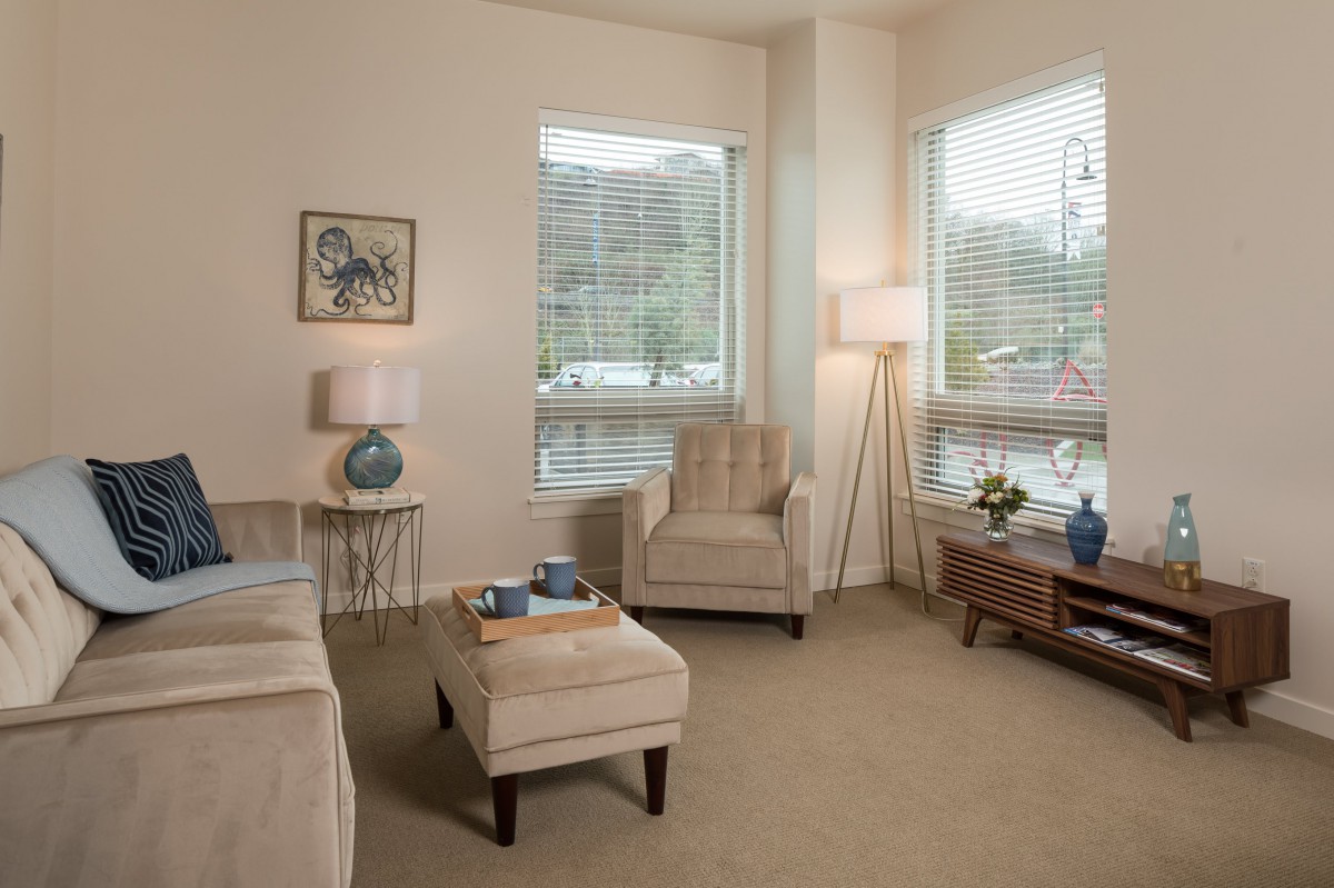 A cozy and modern living room with neutral tones, featuring a plush armchair, ottoman, and large windows allowing ample natural light.