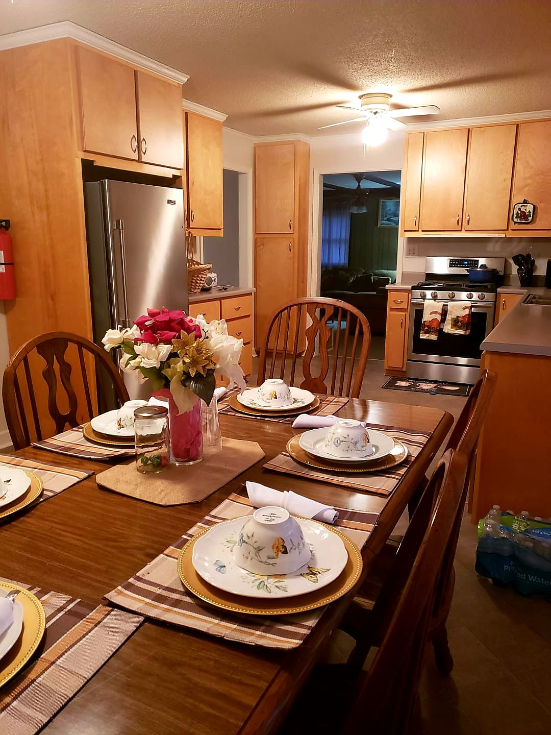 A cozy kitchen with a wooden dining table set for six, featuring floral decorations and surrounded by wooden cabinets and modern appliances.