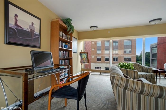 A cozy living room with a home office setup, featuring a computer desk and bookshelf alongside large windows.