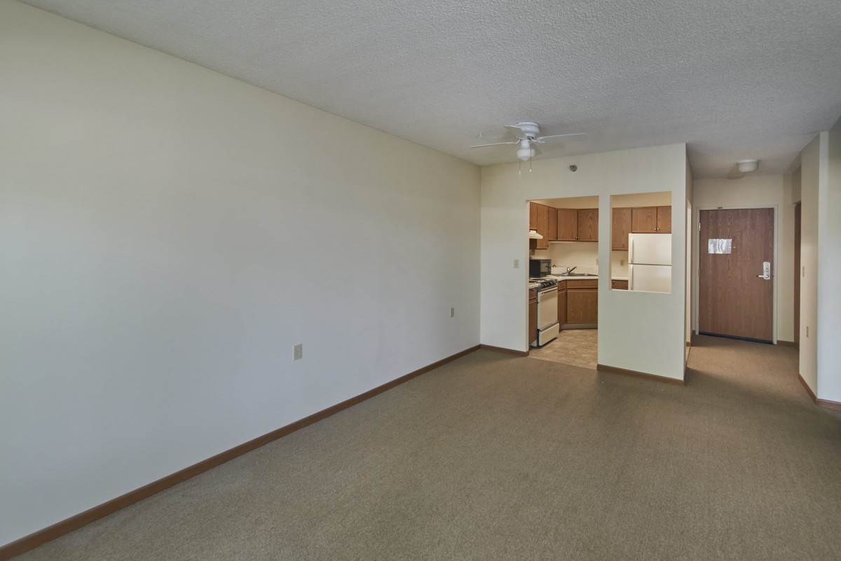 A spacious empty room with a carpeted floor and a ceiling fan, offering a view into the kitchen area.