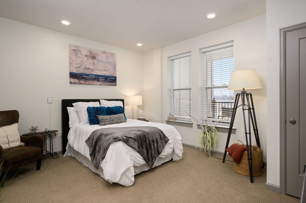 A cozy bedroom features a neatly made bed, modern decor, and ample natural light from large windows.